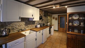 Una cocina con armarios blancos, pared de piedra, vigas de madera, un lavamanos, un microondas, un lavavajillas y un mueble con vajilla.