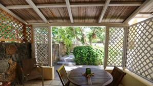 Um alpendre coberto com uma mesa redonda e cadeiras, uma parede de pedra e vista para um jardim através de uma porta ajourada.
