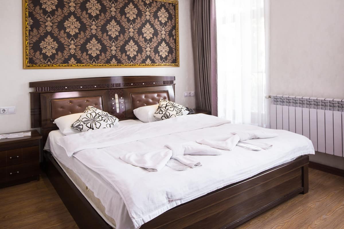 A neatly made bed with white linens, a large patterned tapestry above the headboard, and a window with curtains.