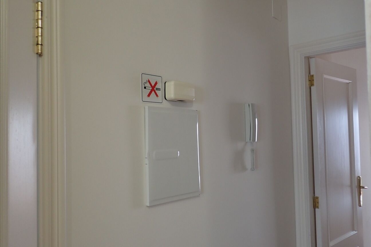 Una puerta blanca con manija de latón, un portero automático instalado en la pared y un detector de humo.