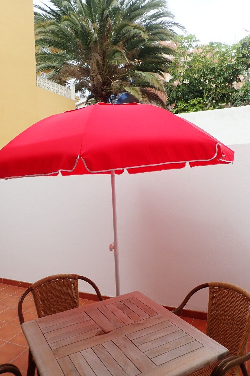 Een terras met een tafel, stoelen, een rode parasol en een palmboom op de achtergrond.