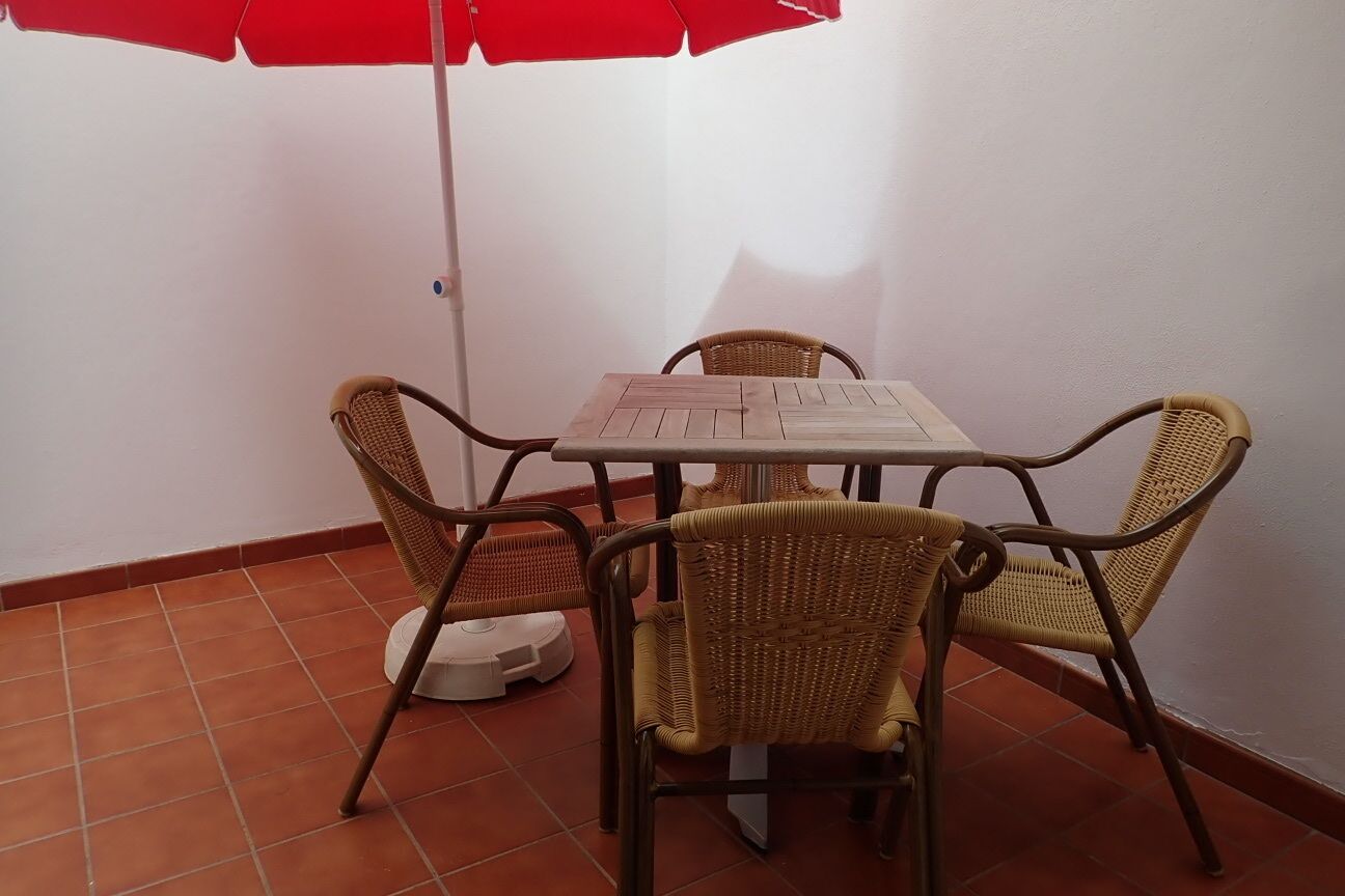 Un petit espace extérieur aménagé pour les repas, avec une table et quatre chaises en osier, un parasol rouge et une lampe de sol blanche.