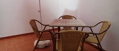 Un petit espace extérieur aménagé pour les repas, avec une table et quatre chaises en osier, un parasol rouge et une lampe de sol blanche.