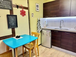Een kleine keuken met een blauwe tafel, houten stoelen, een koelkast en donkere houten kastjes.