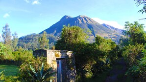 Una montaña bajo un cielo despejado, rodeada de vegetación y un sendero que conduce a una estructura de piedra.