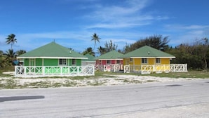 Une rangée de charmantes maisons de plage aux couleurs vives, dotées de balustrades blanches et d’un toit vert, se dressent sur une plage de sable fin, avec des palmiers en arrière-plan.