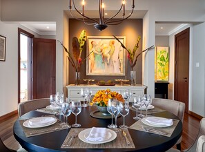 Un coin repas avec une table ronde dressée pour six personnes, entourée de chaises. On y trouve deux vases ornés de branches décoratives et un arrangement floral central composé de fleurs oranges. La pièce est agrémentée d’un lustre, d’un tableau au mur et d’une porte située à gauche.