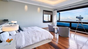 Una habitación de hotel moderna con una cama grande, un escritorio con lámpara, una silla, un telescopio y un ventanal con vista al océano.