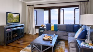 Una sala moderna con un amplio sofá modular gris, un televisor de pantalla plana, una mesa de centro de madera y vista al océano a través de amplias ventanas.