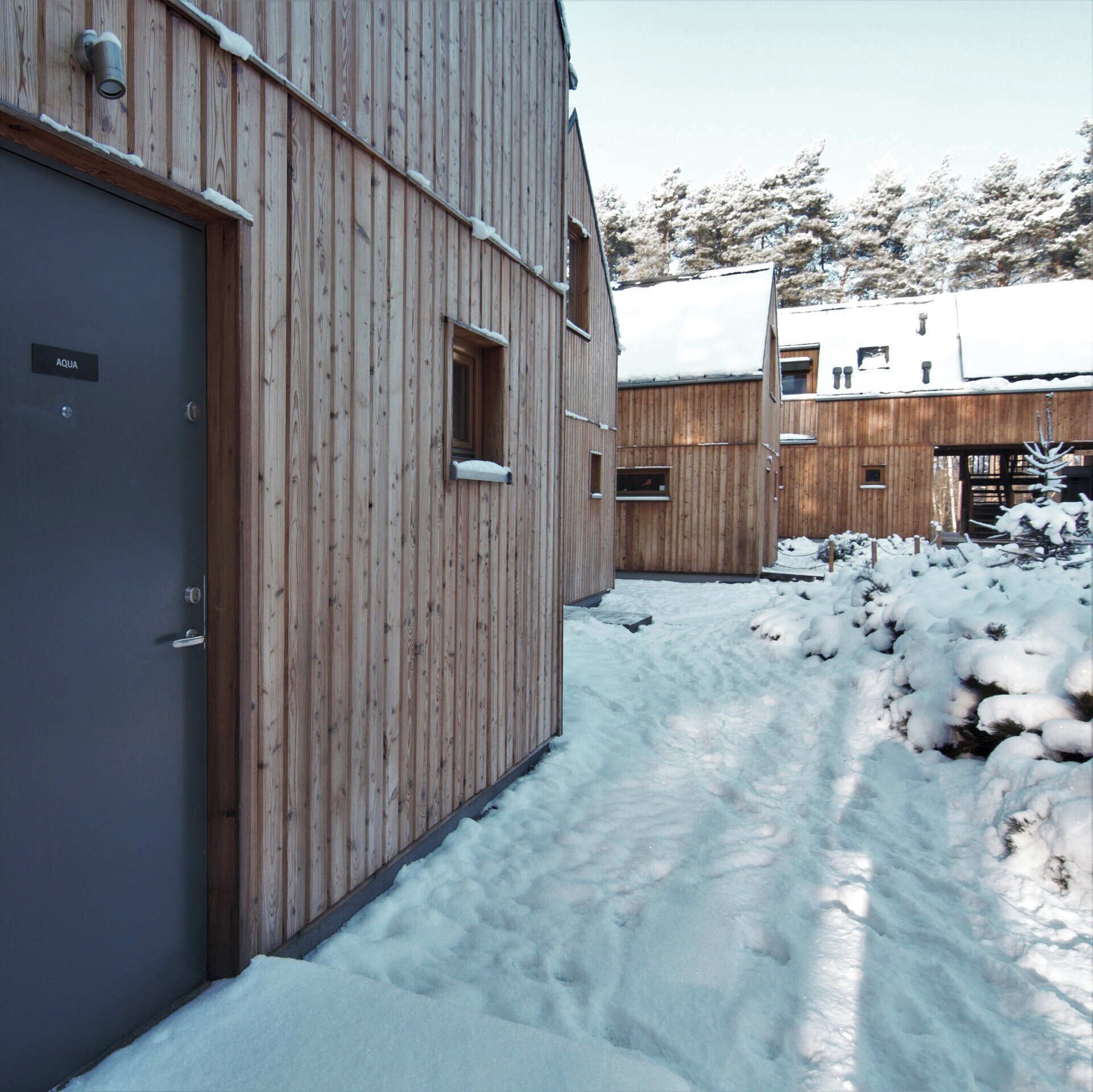 Un edificio de madera con una puerta que dice "AQUA" y una ventana. La nieve cubre el suelo y los árboles al fondo.