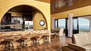 Amplia sala de estar con cocina, taburetes de bar, sofá y ventanas grandes con vistas.
