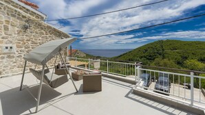 Un balcon avec un fauteuil suspendu, des meubles en osier et une vue sur une colline verdoyante et la mer.