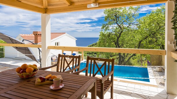 Une terrasse en bois avec une table dressée pour le petit-déjeuner, d’où l’on peut admirer une piscine et une vue sur la mer.