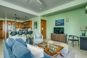 A modern living room with a sofa, coffee table, and kitchen area with wooden cabinets and a TV. - 3BR Condo 421: Dual Balconies, Sea View,Sleeps 6 (Seine Bight)