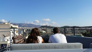 Un couple est assis sur un balcon, d’où l’on peut admirer une vue sur la ville, avec ses bâtiments et une colline qui se dessine à l’horizon.