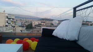 Un balcon avec un canapé noir et des balles colorées, d’où l’on peut admirer un paysage urbain avec des bâtiments et des montagnes.
