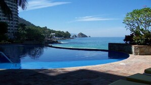 Una piscina con borde curvo, vista despejada del océano y un edificio con balcones al fondo.