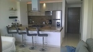 Una cocina moderna con una isla central, taburetes de bar, electrodomésticos de acero inoxidable y una zona de comedor con una mesa y sillas blancas.
