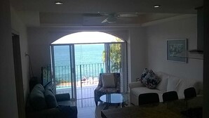 Una sala de estar con un sofá, un sillón y una mesa de centro. Hay un balcón con vistas al océano.