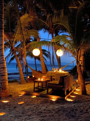 Een eettentje aan het strand met een tafel en stoelen, omringd door palmbomen en uitzicht op de oceaan.