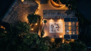 Vue aérienne d’un complexe hôtelier situé en bord de mer, avec une structure à toit de chaume, un espace détente avec des coussins et des palmiers.