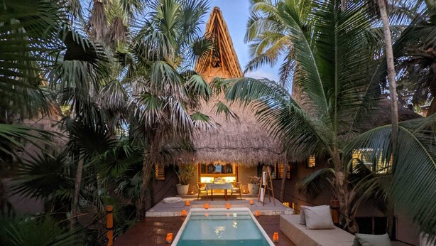 Un espace piscine agrémenté d’une structure à toit de chaume, entouré de palmiers et doté d’une terrasse en bois.