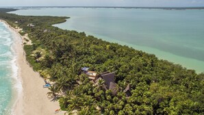 Vue aérienne d’un complexe hôtelier en bord de mer, avec un grand bâtiment entouré d’une végétation tropicale luxuriante et une longue étendue de plage.
