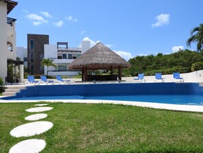 En swimmingpool med blå fliser, omgivet af hvide liggestole og en overdækket pavillon med stråtag.