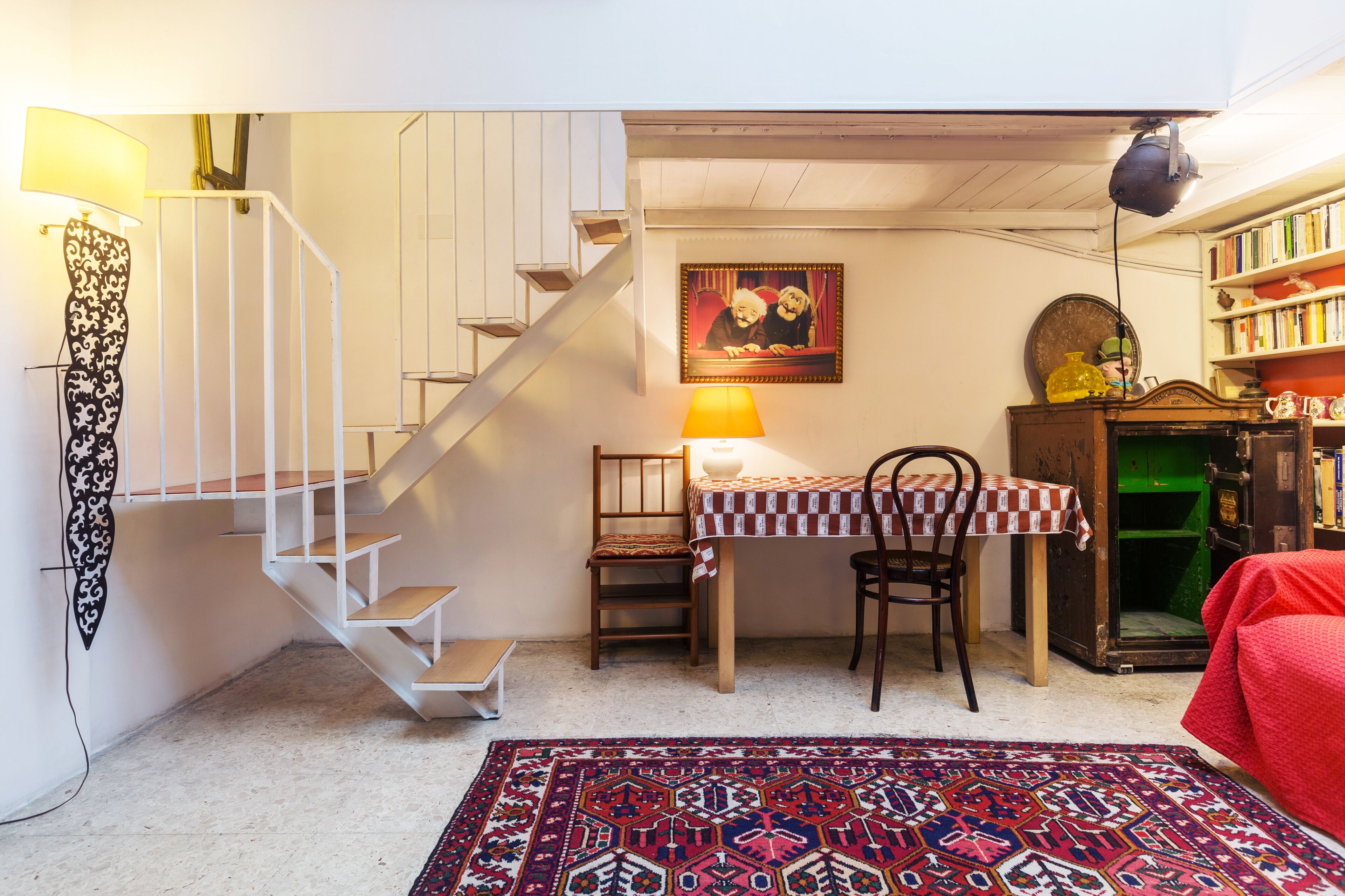 A staircase leading to a second floor, a table with a checkered tablecloth, a chair, a lamp, a bookshelf, and a patterned rug.
