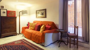 A cozy living room with a sofa, a wooden chair, a small table, a lamp, and a window with curtains.