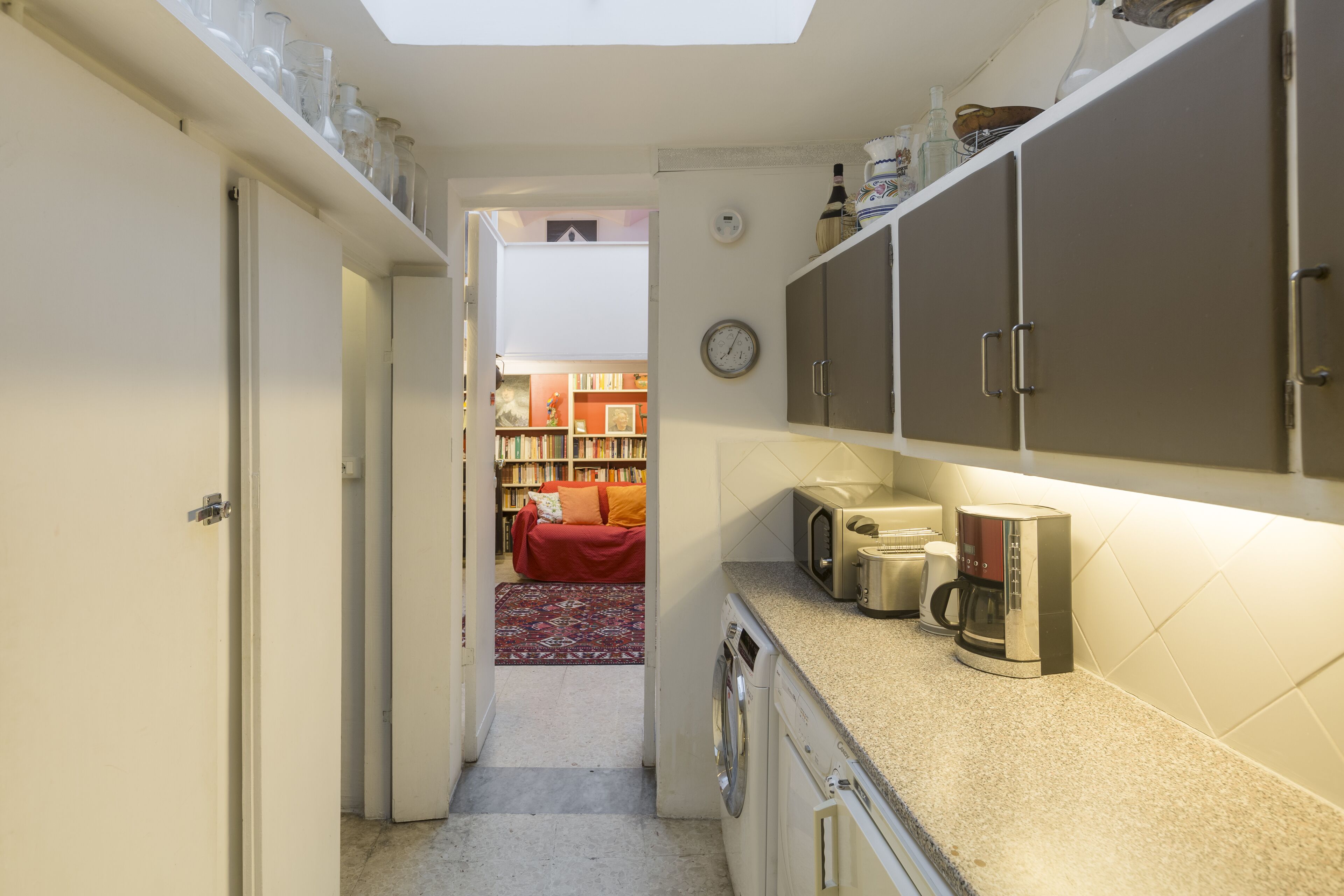 Une cuisine compacte équipée d’un lave-linge, d’un four à micro-ondes et d’une machine à café. On aperçoit une bibliothèque et un canapé rouge à l’arrière-plan.