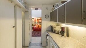 A compact kitchen with a washing machine, a microwave, and a coffee maker. A bookshelf and a red sofa are visible in the background.
