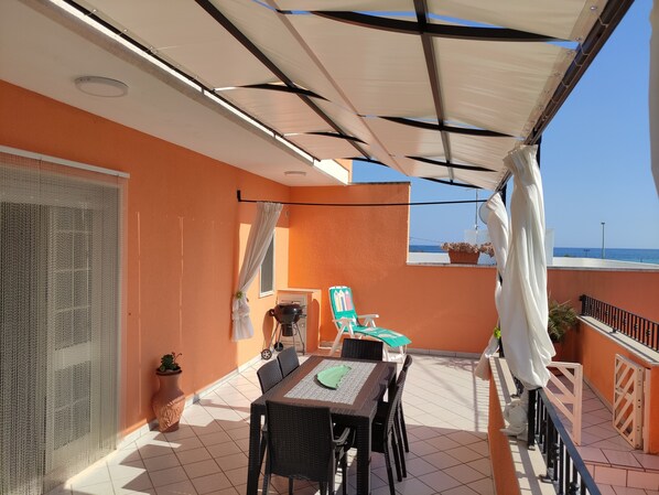 A balcony with a table and chairs, a grill, and a view of the sea.