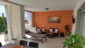 A sunlit patio with a dining table, chairs, a cushioned seating area, and potted plants.