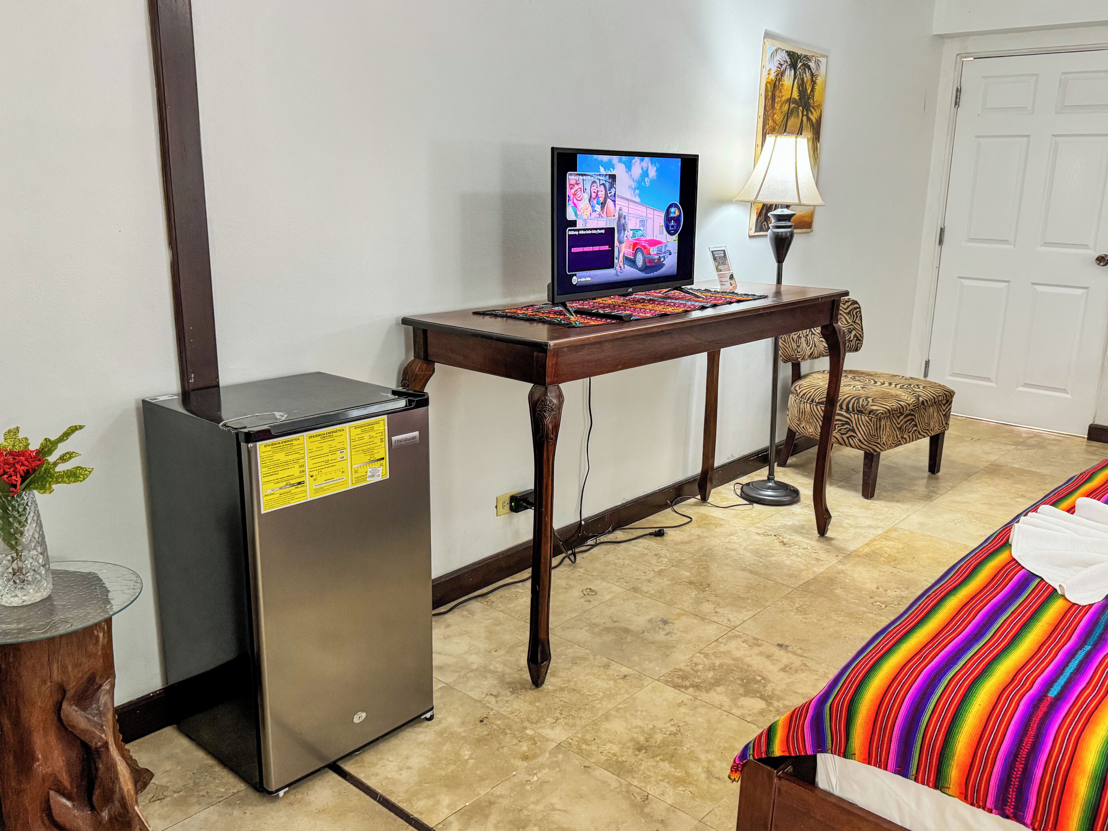 A compact hotel room with a bed, a desk with a computer, a refrigerator, and a small table with a lamp.