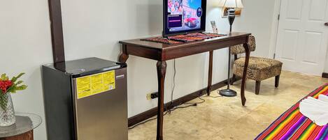 A compact hotel room with a bed, a desk with a computer, a refrigerator, and a small table with a lamp.