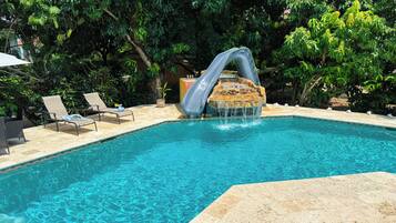 Una piscina con tobogán de agua, sillas de descanso y exuberante vegetación.