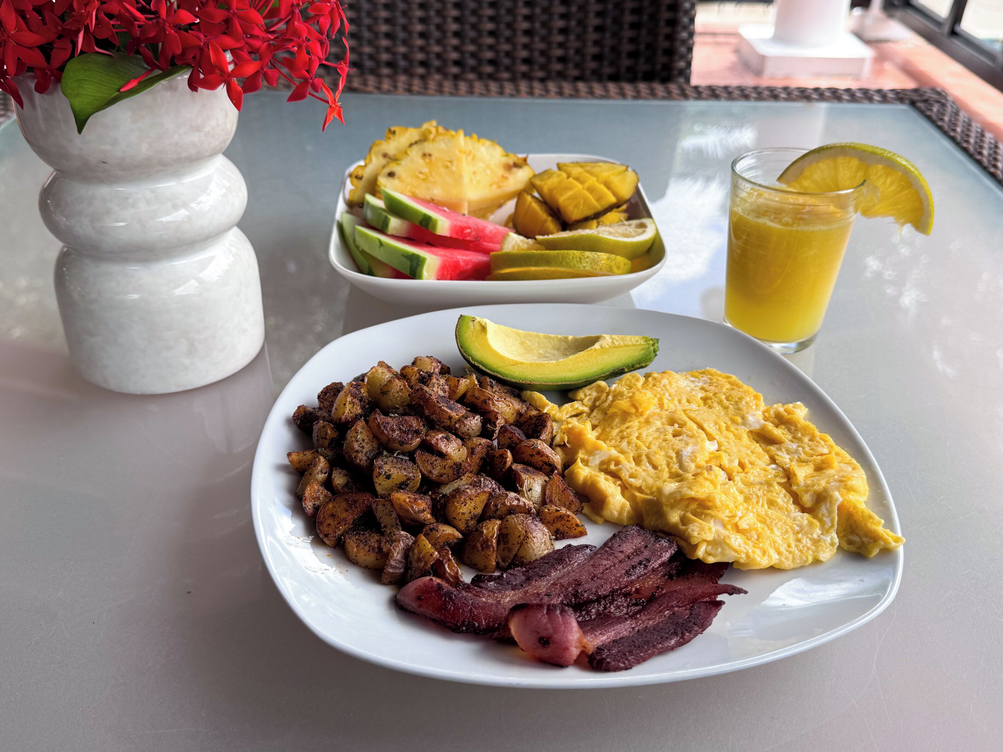 A breakfast plate with eggs, bacon, potatoes, avocado, and a fruit bowl with watermelon and mangoes.