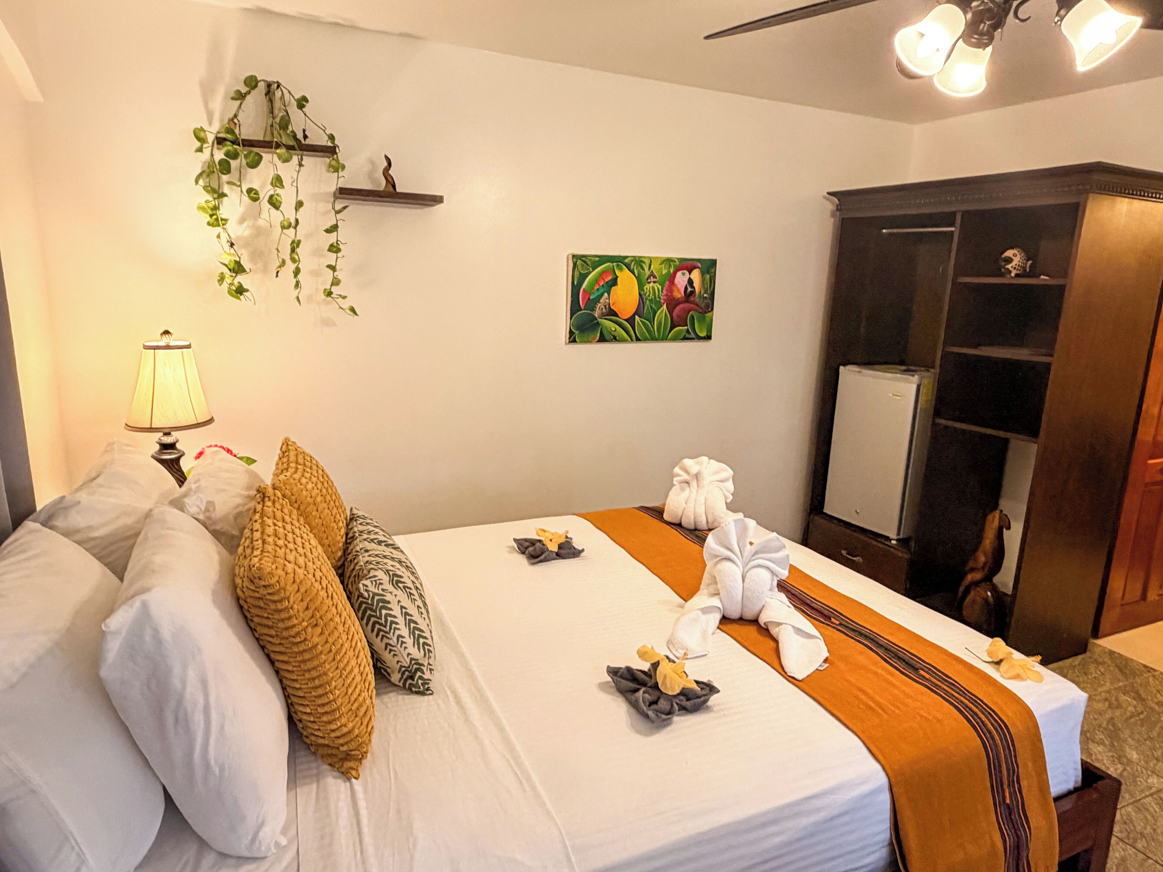 Ein Hotelzimmer mit Bett, Kissen, einer Lampe, einem Gemälde, einem Kleiderschrank und einer Pflanze.