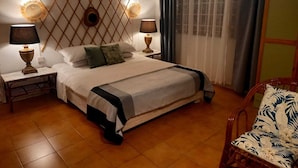 Una cama bien hecha con cabecera, dos mesitas de noche con lámparas, una silla con estampado floral y una ventana con cortinas.