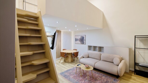 Een modern loft appartement met een houten trap, een ronde eettafel, een beige bank en een vloerkleed met dessin.