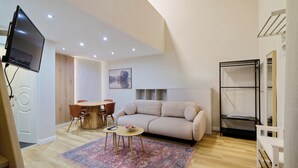 Een moderne woonkamer met een bank, een eettafel en stoelen. Er is een televisie aan de muur bevestigd, een klein vloerkleed en een plank met een spiegel.