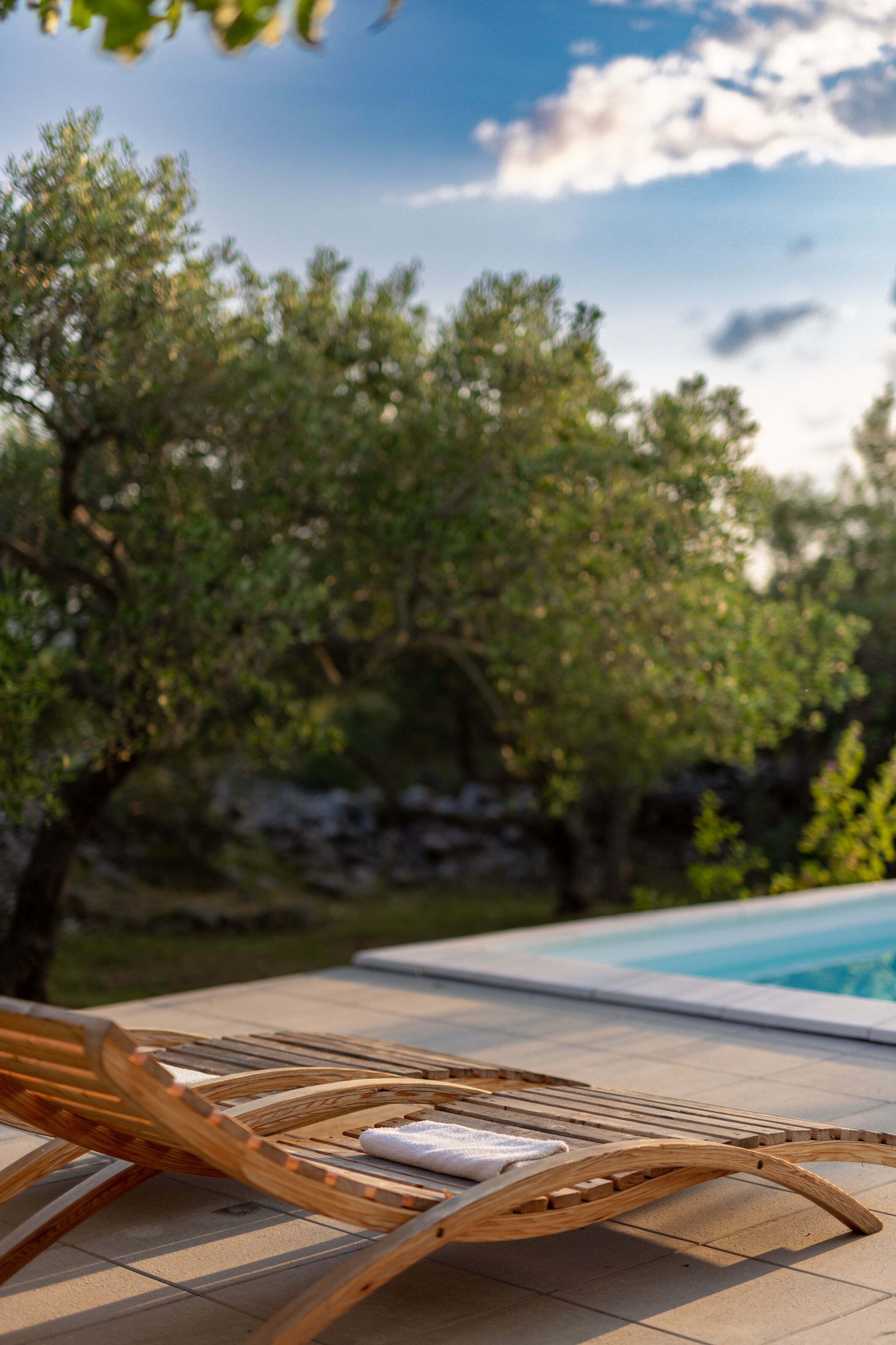 Una poltrona da relax in legno con un asciugamano, una piscina e alberi sullo sfondo.