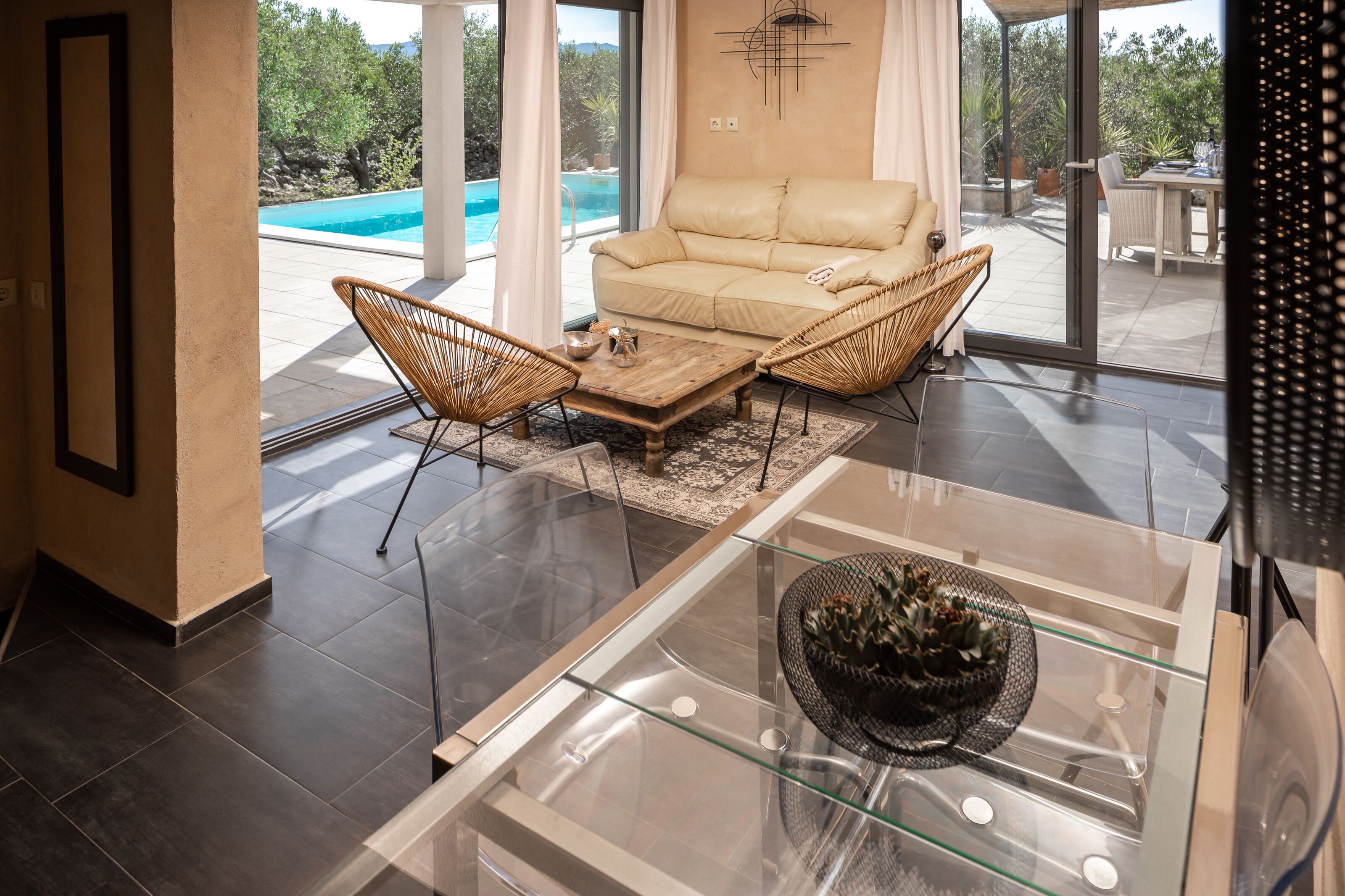 Un salon moderne avec un canapé beige, des fauteuils en osier, une table basse en verre et une vue sur une piscine.