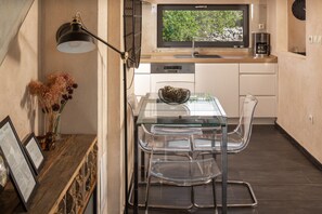 A modern kitchen with a glass dining table, clear chairs, a black pendant light, and a window with a view of greenery.