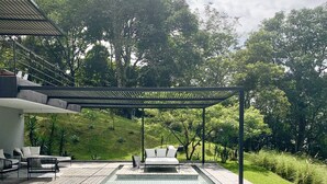 A rectangular swimming pool with a pergola overhead, surrounded by a paved deck and lounge chairs.