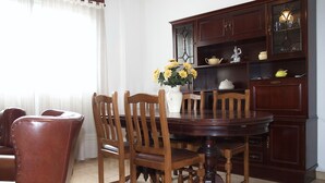 Un comedor con una mesa redonda de madera, seis sillas de madera, un jarrón blanco con flores amarillas y un aparador de madera oscura con puertas de vidrio.