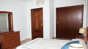 Un dormitorio con cama, cómoda, espejo, dos puertas y mesita de noche con lámpara.