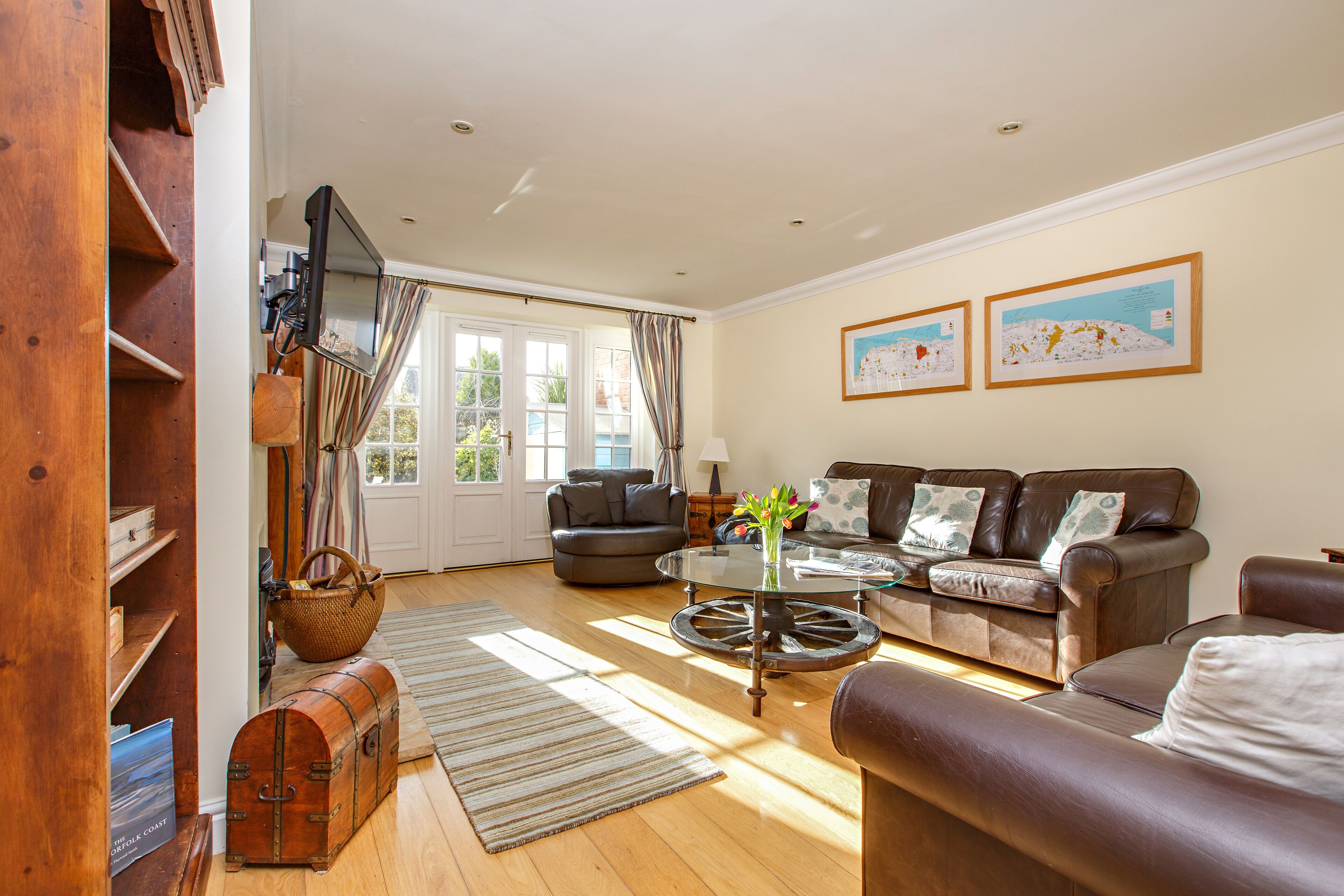 A living room with a brown leather sofa, a glass coffee table, and a television mounted on the wall. There are two framed pictures on the wall and a window with curtains.