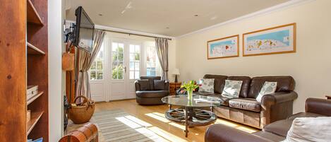 A living room with a brown leather sofa, a glass coffee table, and a television mounted on the wall. There are two framed pictures on the wall and a window with curtains.
